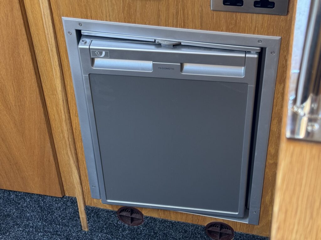 A gray under-counter refrigerator installed in a wooden cabinet, featuring a silver handle and a sleek design. The cabinet has a light-colored wood finish, and the floor is covered with dark carpet.