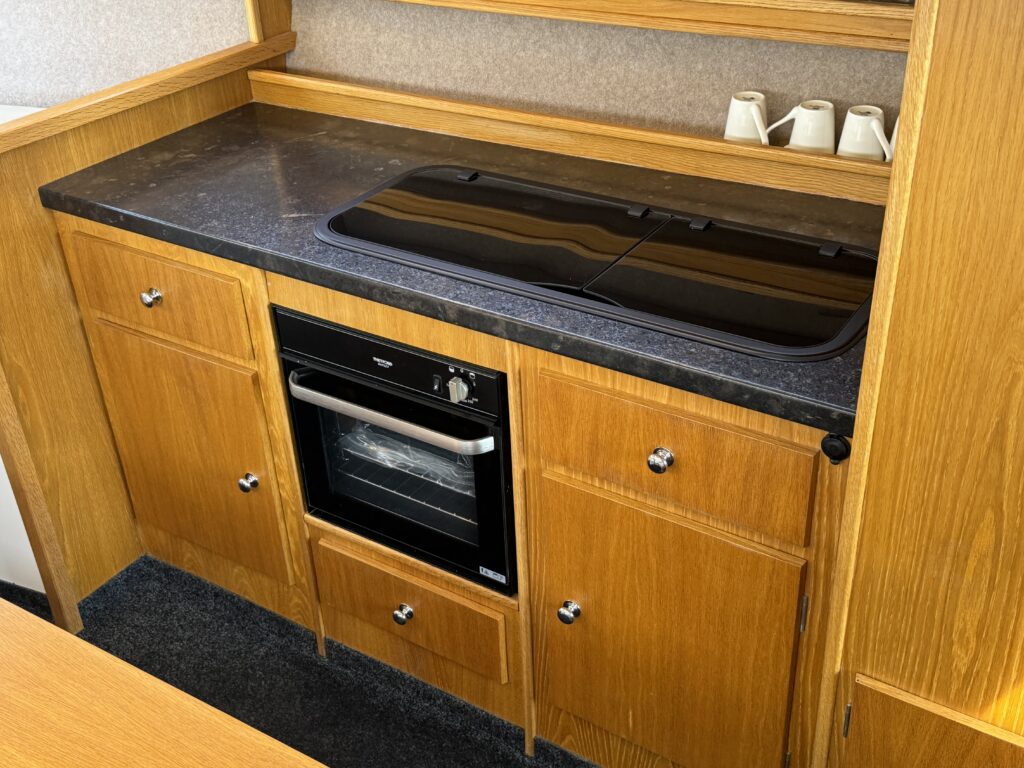 A wooden kitchen cabinetry featuring a black countertop with a cooktop and an oven below. The lower section has two drawers and two cabinet doors, with metallic handles. A few white cups are visible on a shelf in the background.