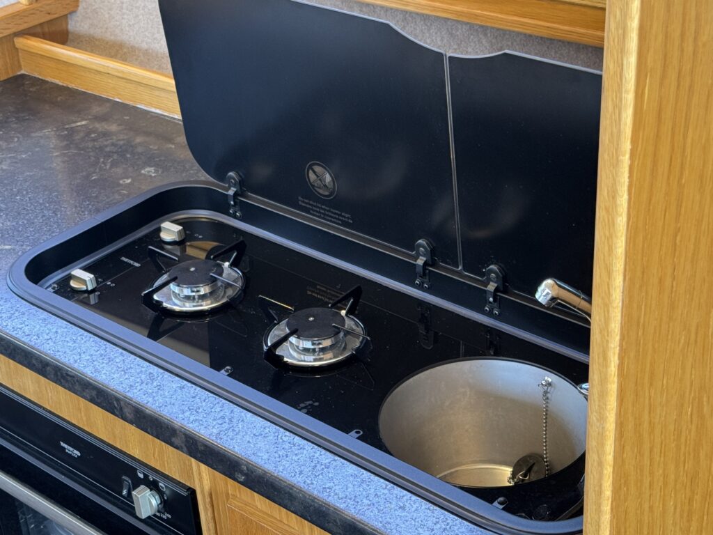 A black gas stove with two burners is mounted on a countertop. The stove has a hinged cover, slightly open, revealing a metal basin beside the burners. The countertop is a blue granite-like surface, and there is wooden cabinetry above.