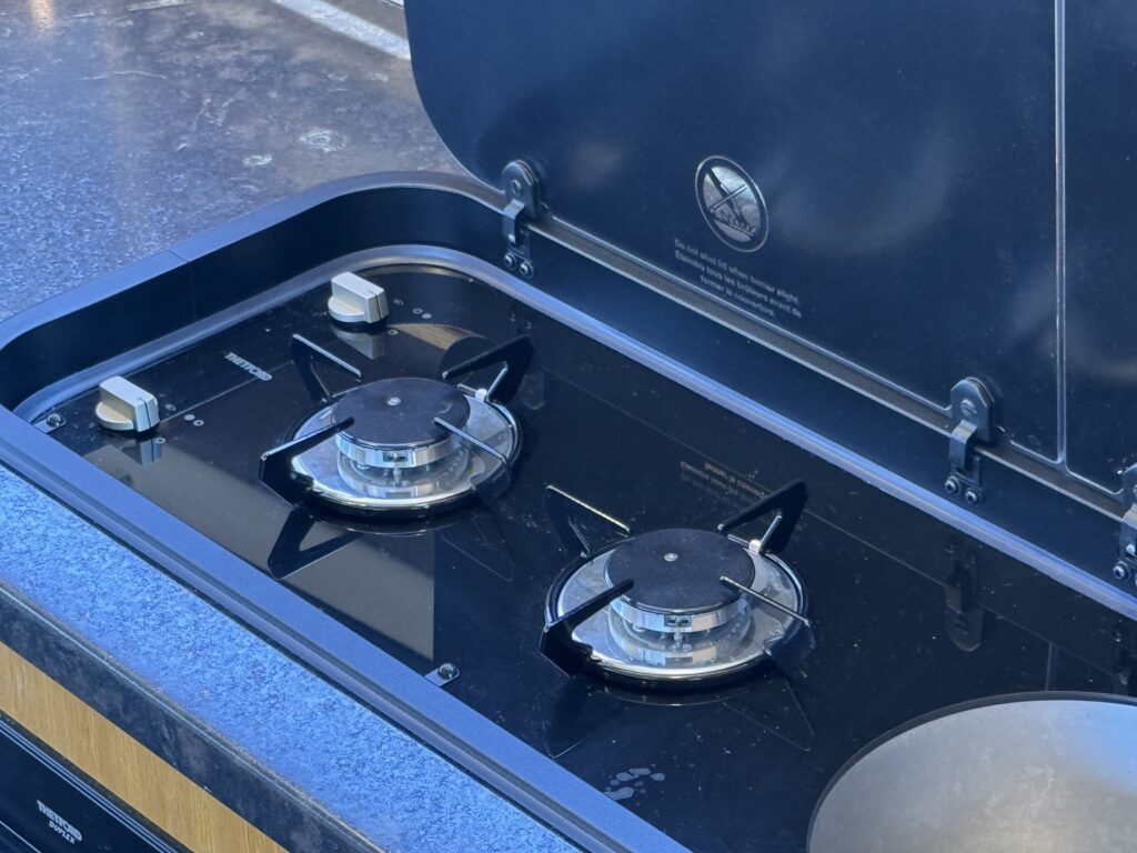 A gas cooktop with two burners featuring black knobs and stainless steel grates, set into a countertop. The lid is open, revealing the glossy black surface with a warning label above the burners.