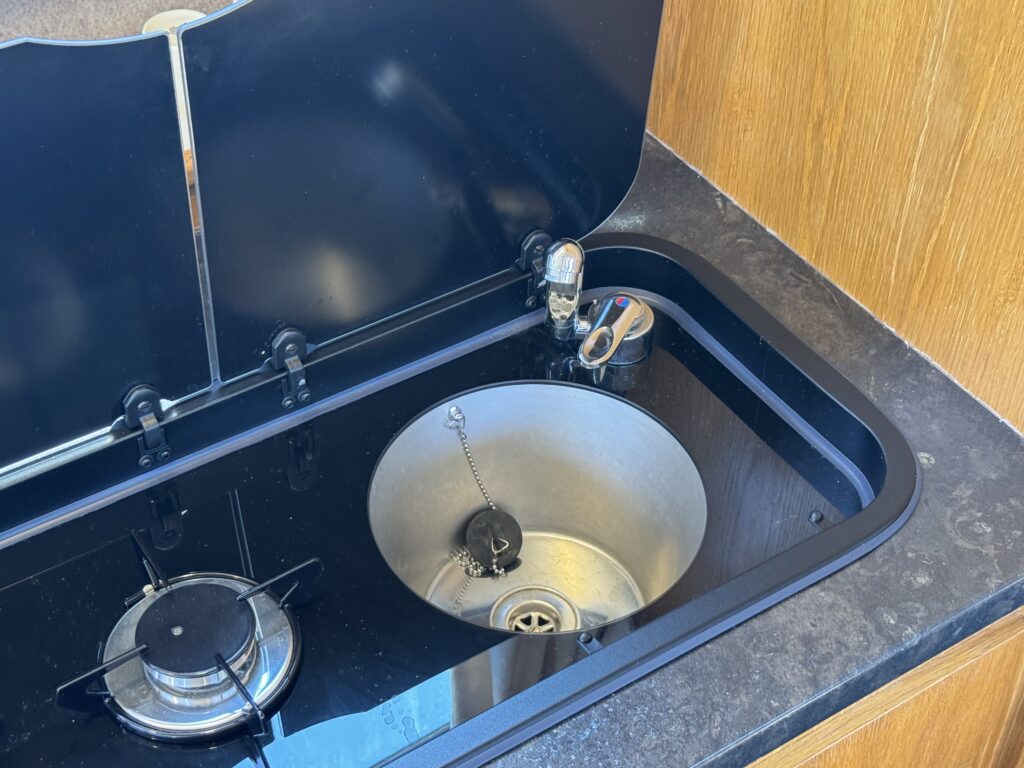 A black sink with a circular basin and a visible drain is installed on a countertop. To the left, there is a small gas burner. The countertop has a wooden backing, and the sink lid is closed, revealing a minimalist design.