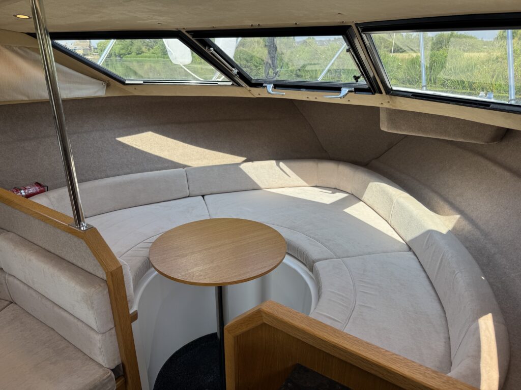 An interior view of a boat featuring a circular seating area with beige cushions and a wooden table in the center. Large windows allow natural light, illuminating the space. The walls are lined with light-colored material, creating a cozy atmosphere.
