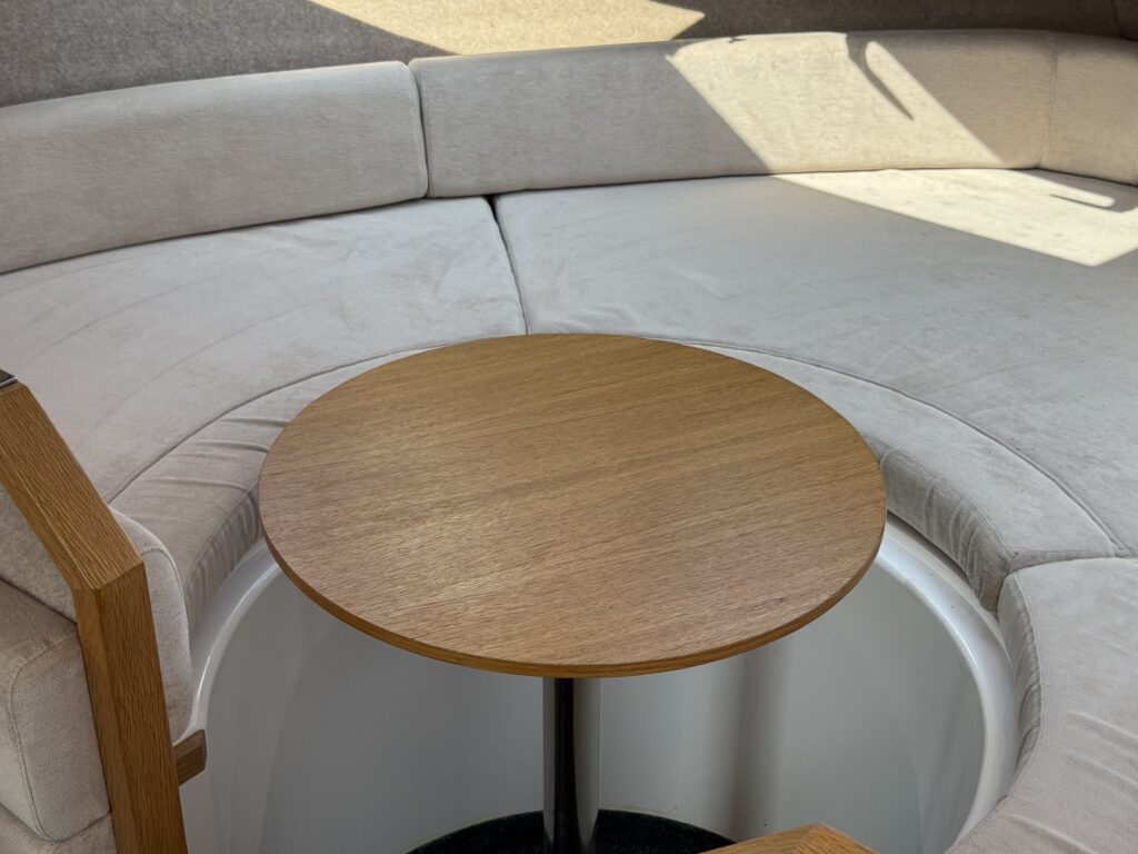 A circular wooden table stands in the center of a seating area with a beige cushioned banquette. The seating features a curved design, and natural light illuminates the space from above, highlighting the comfortable materials.