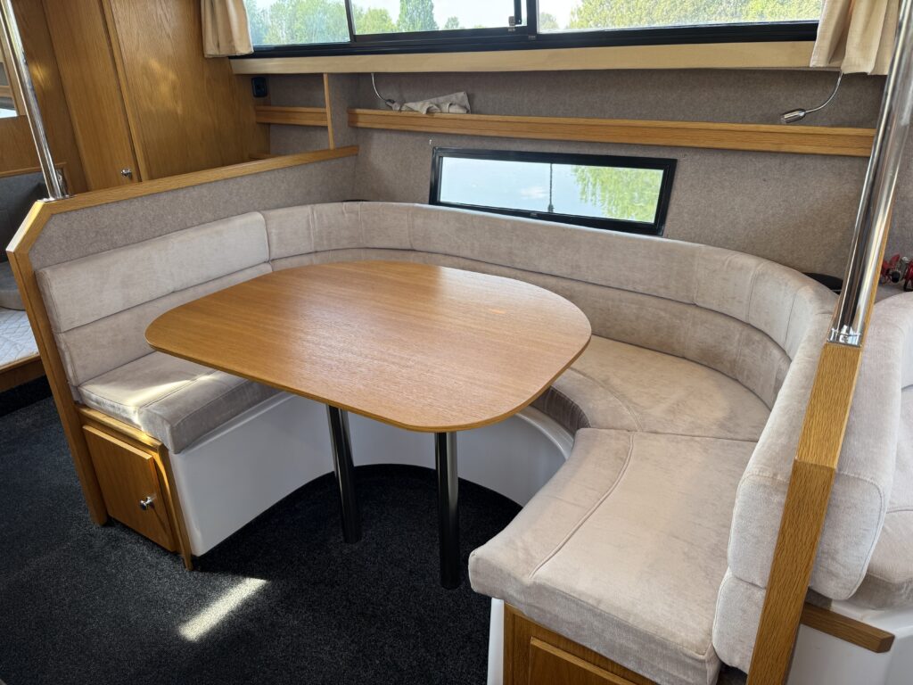 A cozy dining area featuring a rounded, light-colored upholstered bench, a wooden oval table, and dark carpet. Large windows provide natural light, and shelves are installed along the walls.
