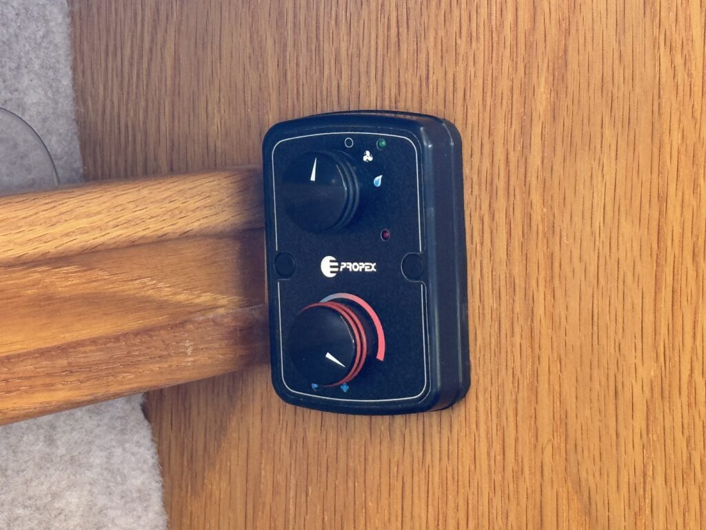 A black control panel with two circular knobs is mounted on a wooden surface. One knob is labeled with a red accent, and the other has a blue indicator, with small lights and symbols above them.