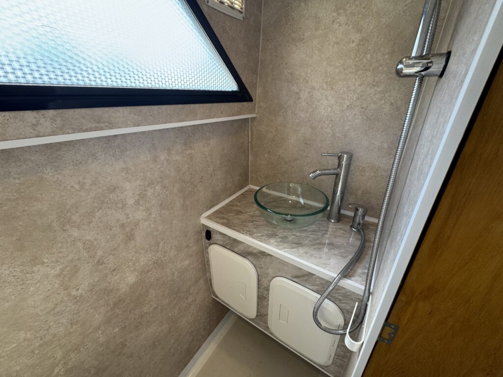 A small bathroom area features a glass vessel sink on a marble countertop with a modern faucet. The wall has a window and a showerhead is attached nearby. The decor includes neutral tones with a textured finish.