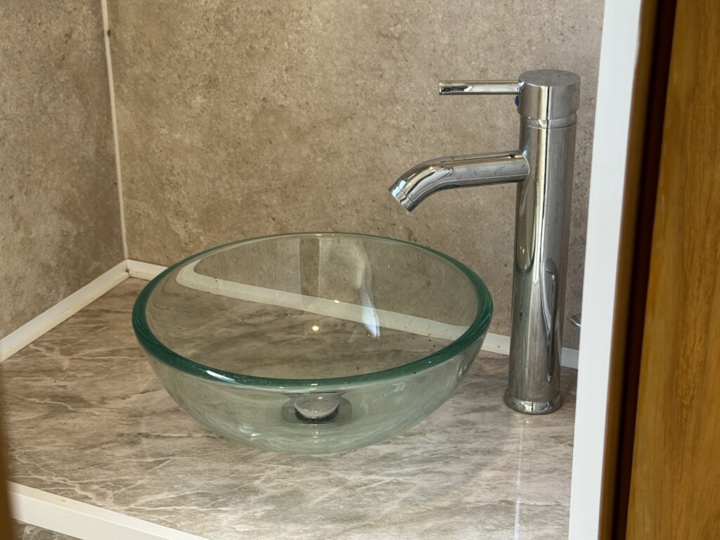 A clear glass vessel sink with a chrome faucet is positioned on a light-colored marble countertop. The wall behind the sink is smooth and gray, complementing the modern design of the space.