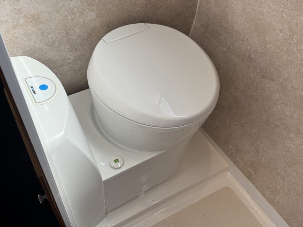 A white compact toilet with a smooth round lid, positioned on a light-colored floor. Next to the toilet, there is a control panel with a blue button and a green indicator light, against a textured beige background.
