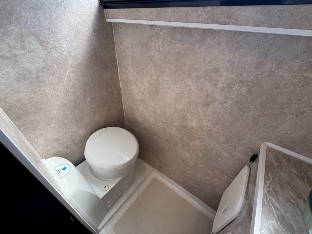 A compact bathroom featuring a white toilet positioned against a textured beige wall. The floor is covered with a light-colored mat, and a small waste bin is visible next to the toilet.