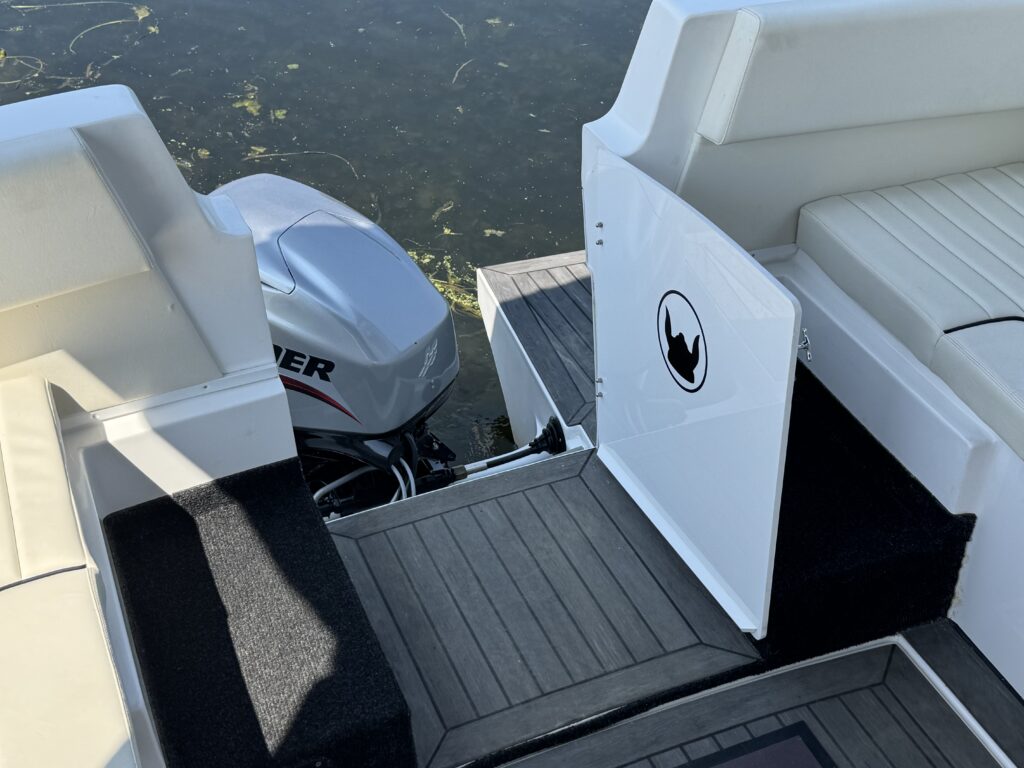 The image shows the corner of a boat featuring a gray outboard motor and a hatch door labeled with a black silhouette of a bird. The deck is made of dark materials, and there are white seating areas visible in the background.