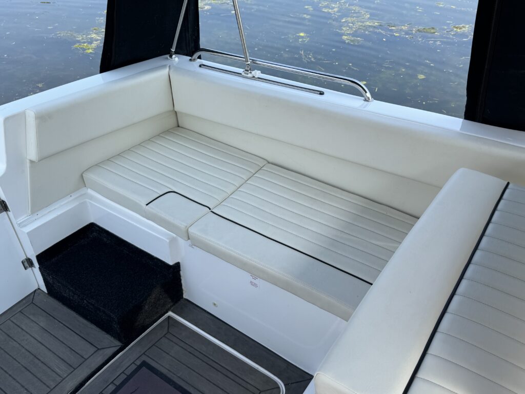 A white cushioned seating area on a boat, with a textured pattern on the cushions. The seating wraps around the corner, and a set of black steps leads down to a lower deck, while the water with green vegetation is visible in the background.