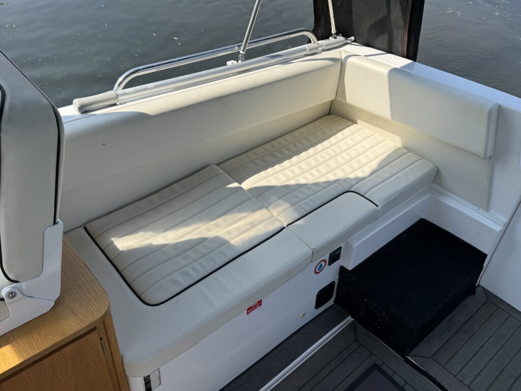 A cushioned seating area on a boat, featuring a light-colored, quilted cushion resting on a white base. The background shows calm water, and the boat's textured flooring is partially visible adjacent to the seating.