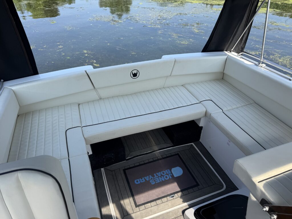 A spacious white boat seating area with a textured cushion design, featuring a center table and open space for movement. The boat is positioned above a calm water surface with greenery reflecting off it, and is partially shaded by a black canopy.