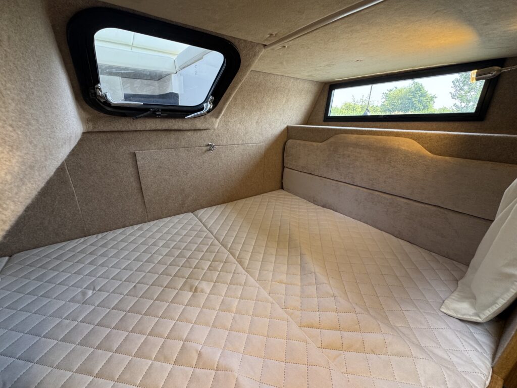An interior view of a small sleeping area with a padded bed covered in a quilted mattress. Two windows allow natural light in, and the walls are lined with beige fabric. The setting appears cozy and compact, designed for comfort in a limited space.