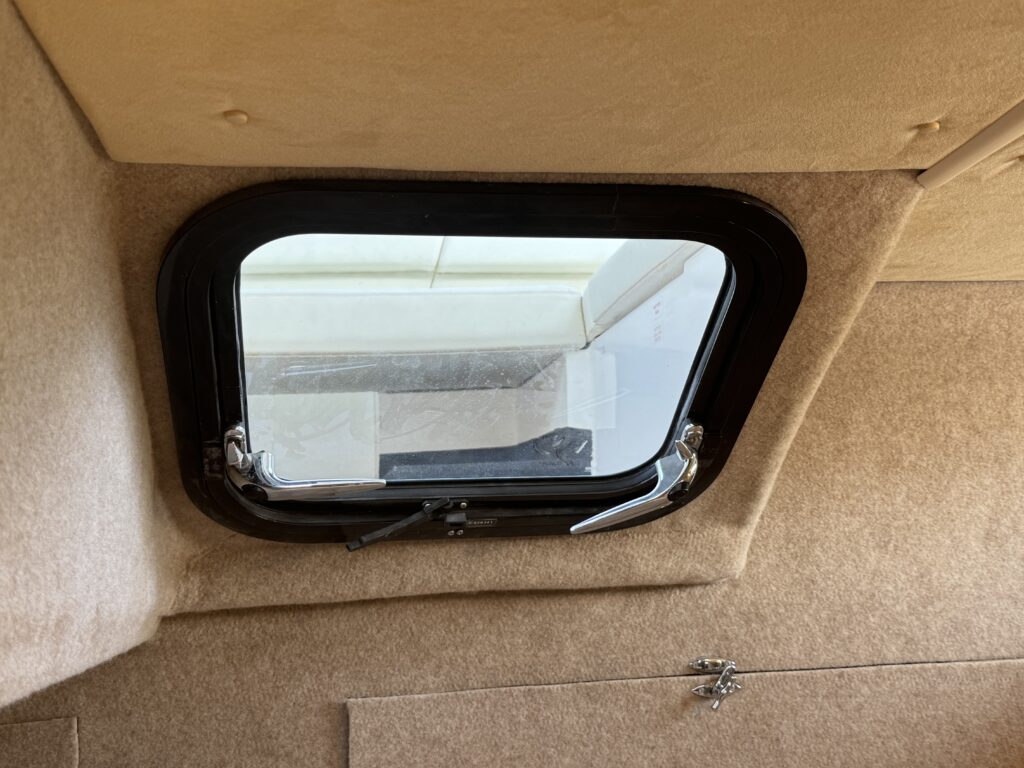 A rectangular window with a black frame is mounted on a tan fabric-covered wall. The window is slightly open, revealing a view of a light-colored surface outside.