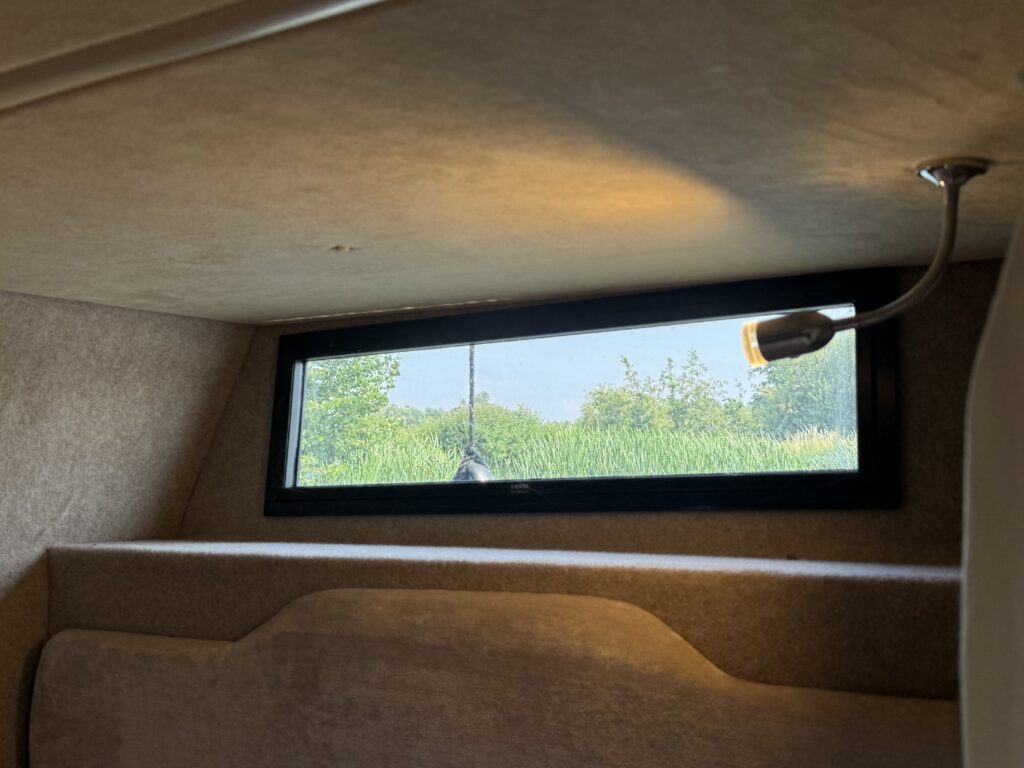 An interior view of a small, cozy space with a window showing green grass and trees outside. A light fixture mounted on the wall is visible, and the walls are covered in a soft, beige material.