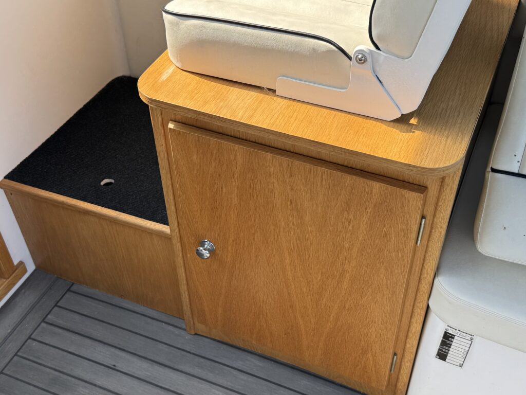 A wooden cabinet with a silver handle, located next to a cushioned seat in a boat. The cabinet is closed and features a texture contrasting with the gray-striped flooring visible underneath.