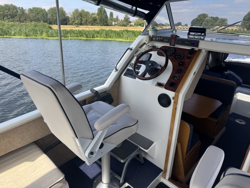 The interior of a motorboat featuring a white captain's chair next to a steering console with various controls, located near a window overlooking a waterway and lush greenery. The floor is dark blue carpet, and there are wooden elements in the design.
