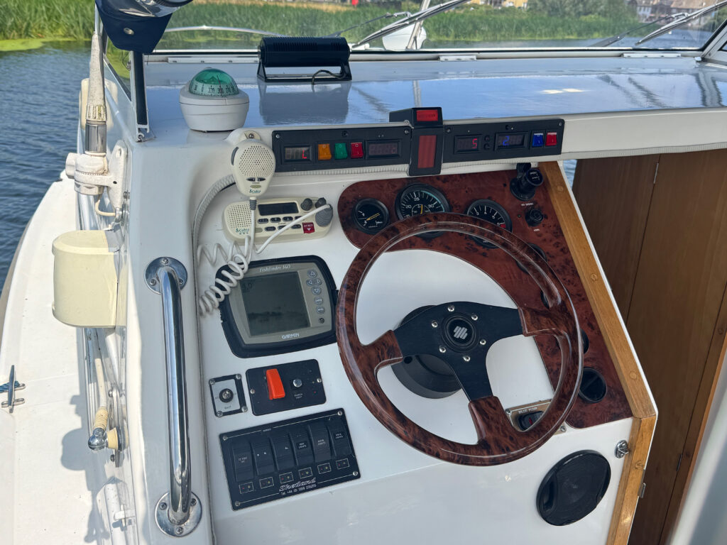 A boat's cockpit featuring a wood and chrome steering wheel, various gauges and controls, a small display screen, and a hand-held radio mounted on the dashboard. The surroundings include water and greenery visible outside.