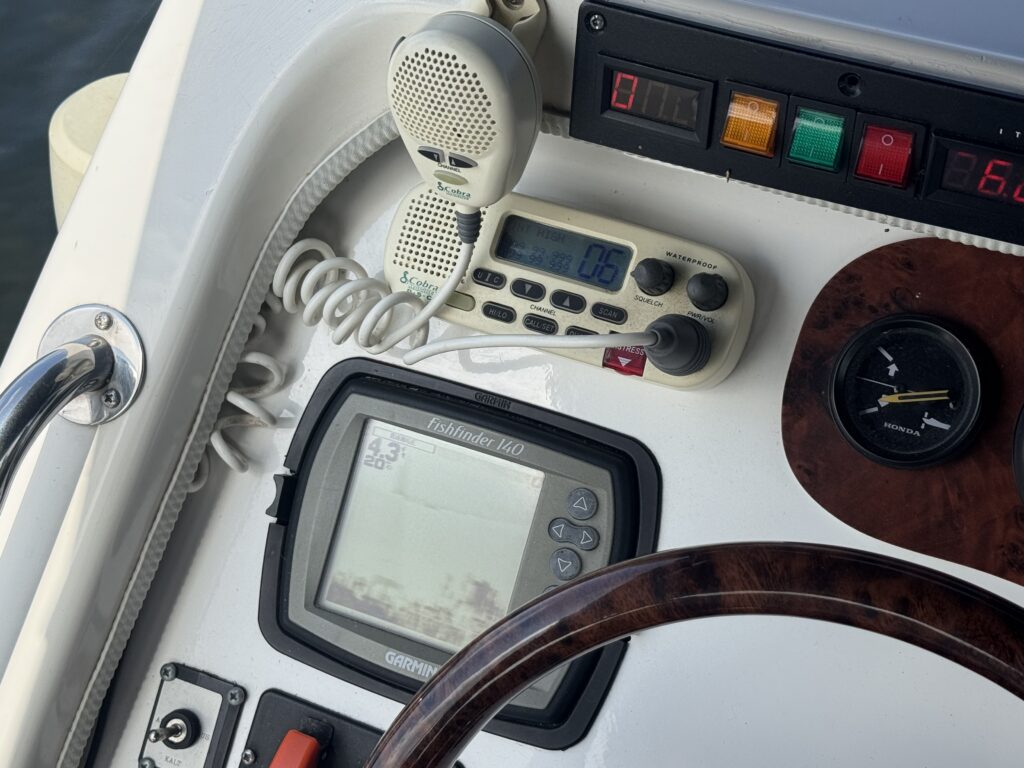 A close-up view of a boat's dashboard featuring a GPS display, a radio communication device, and a set of gauges. The wooden steering wheel is visible, along with buttons and indicators in the control panel.