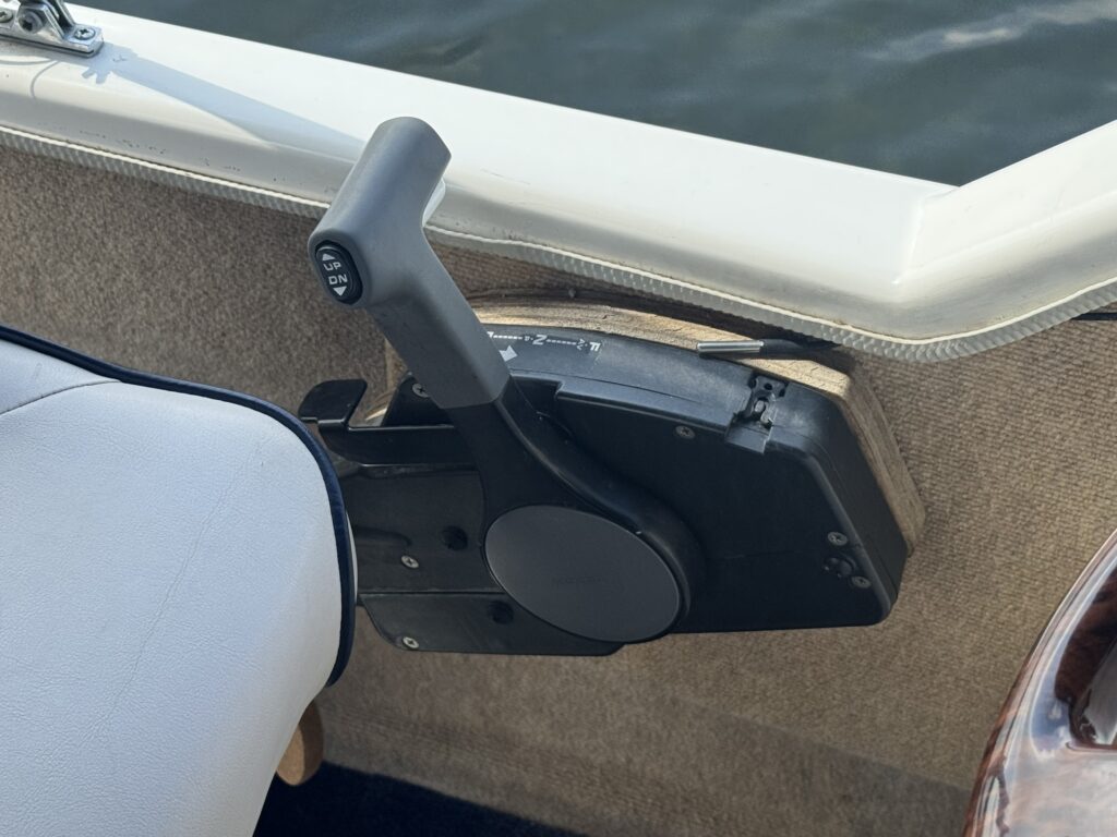 A black throttle lever mounted on the side of a boat, positioned next to a gray upholstered seat. The control is designed for adjusting the boat's speed and direction, with a textured grip and price switches visible on the handle.