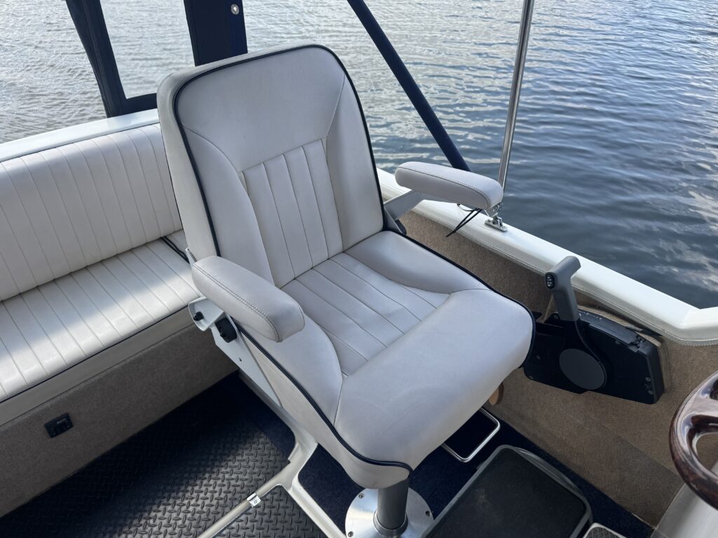 A white upholstered boat seat with vertical stitching and armrests is mounted on a swivel base. The seat is positioned on a deck with dark flooring, overlooking a body of water in the background.