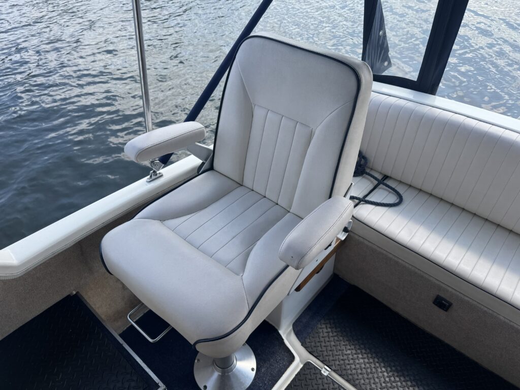 A white padded boat seat with armrests is positioned on a boat deck, with a textured black mat visible underneath. The backdrop features water reflecting sunlight.