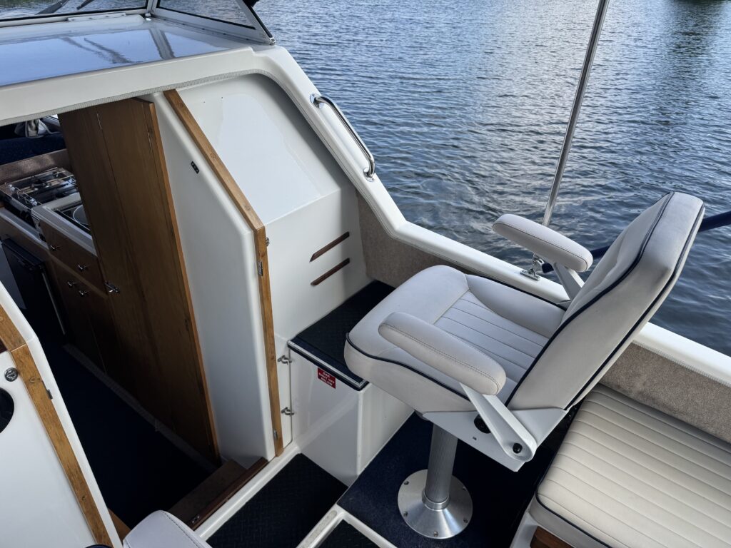 An interior view of a boat showing a gray and white captain's chair mounted on a round metal base. There is a wooden cabinet and door to the left, with a textured mat on the floor and water visible through the windows in the background.