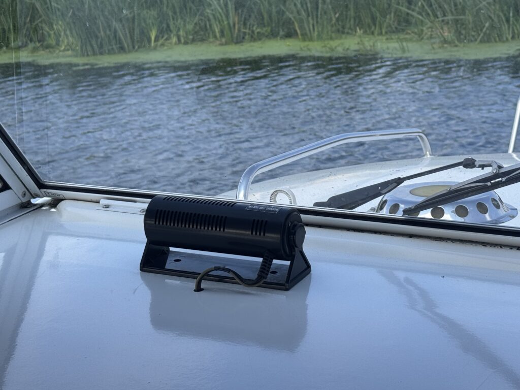 A black boat speaker mounted on a white surface inside a boat, positioned near a window. Outside the window, a view of calm water and green reeds is visible, reflecting natural light.