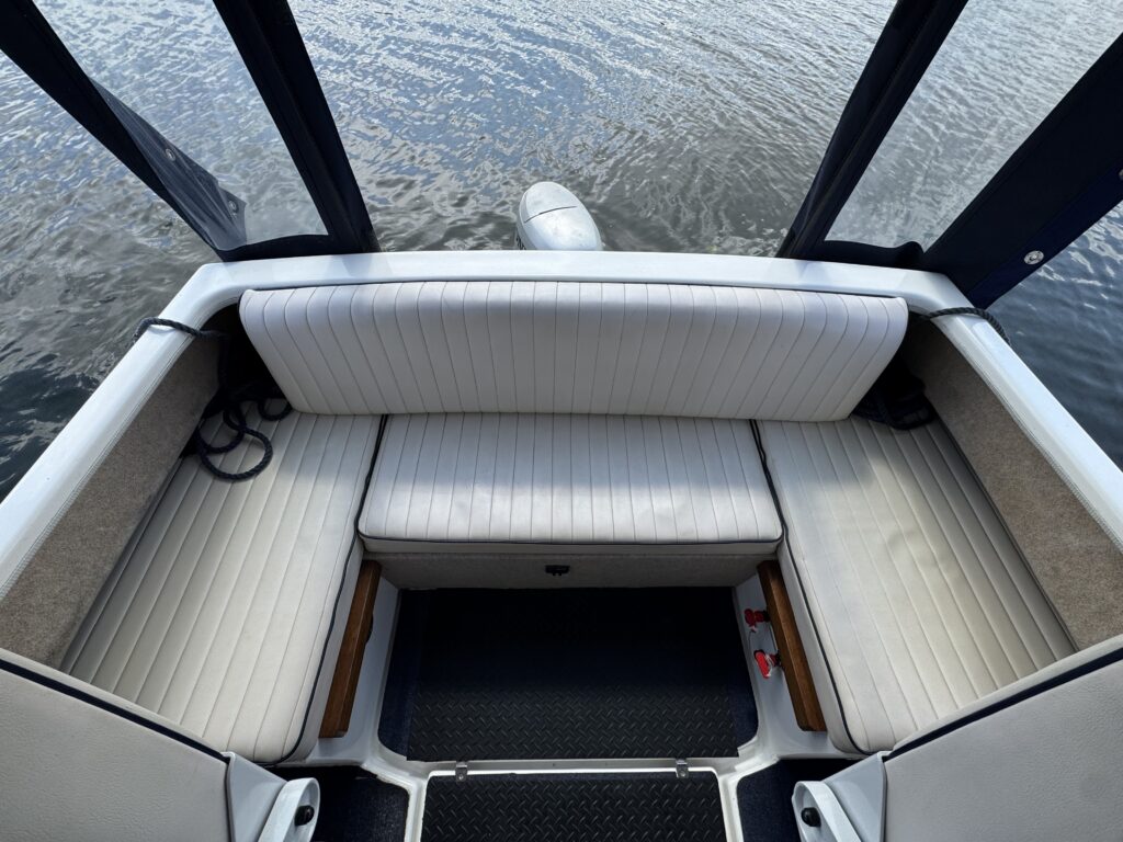 A view of the interior seating area of a boat, featuring white vinyl benches with vertical stripes. The boat is docked on water, and the surface reflects the surrounding area. The seating is arranged in a U-shape, with a flat area in the center.