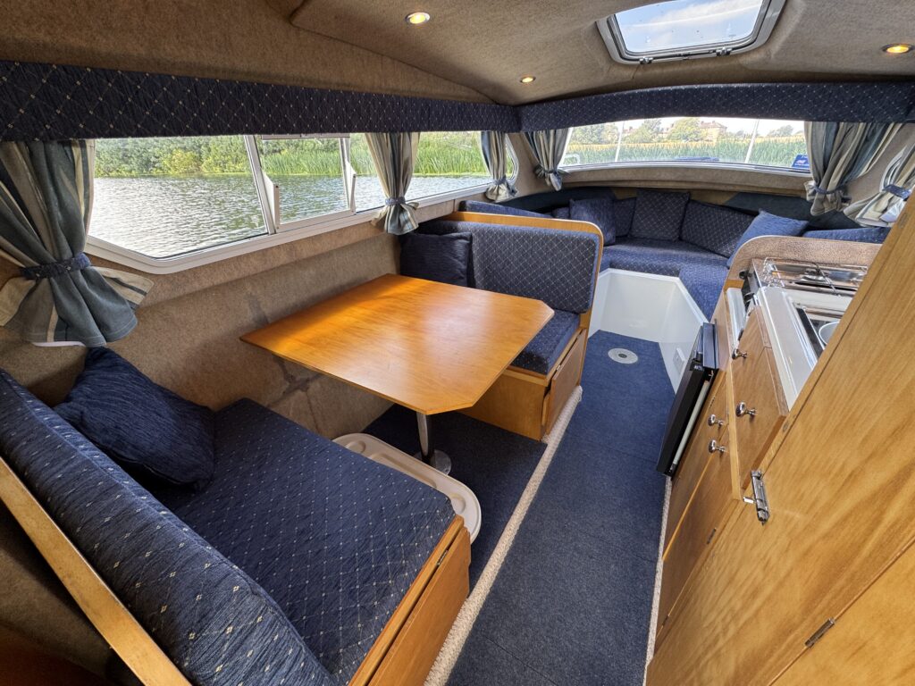 Interior of a boat featuring a spacious lounge area with a wooden table and upholstered seating in blue fabric. The area is well-lit by natural light coming through large windows, and there is a small kitchenette with wooden cabinetry visible to the right.
