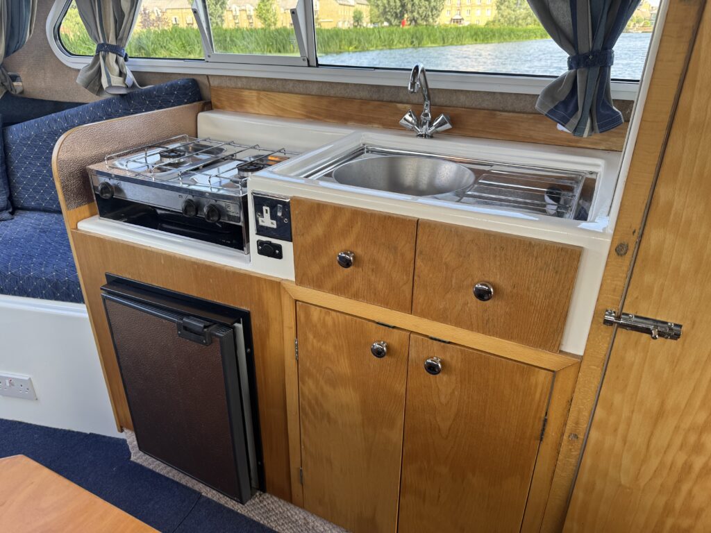 A compact boat kitchen featuring a stainless steel sink, a two-burner gas stove, and a small refrigerator. The cabinetry has a light wood finish, and the area is surrounded by windows with curtains, offering a view of greenery outside.