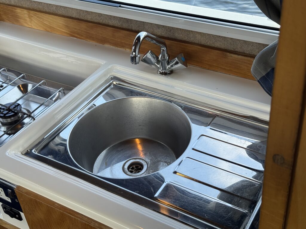 A metal sink in a boat kitchen, featuring a round basin with a drain at the bottom. Next to the sink is a stainless steel countertop with a built-in draining board, and a faucet mounted above. Natural light is visible through a window.