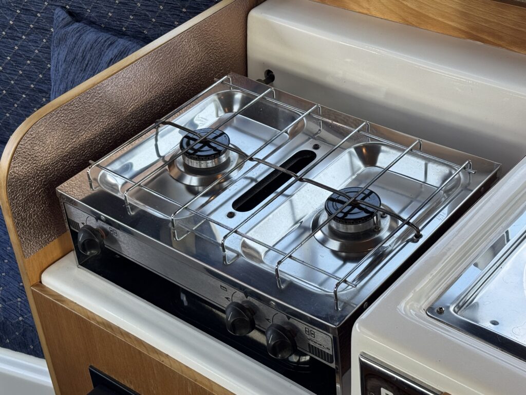 A stainless steel two-burner gas stove with metal grates on a wooden countertop in a compact kitchen space. The stove has two black knobs and a safety port in the center, surrounded by a light-colored surface and a dark blue fabric background.
