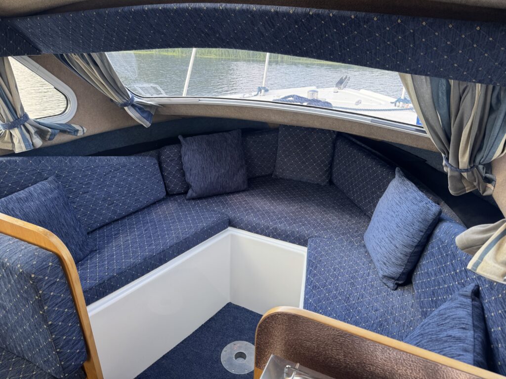 A cozy seating area inside a boat, featuring blue cushioned seats arranged in a semicircular shape. A small white table is centered within the seating, and large windows provide a view of the water outside.