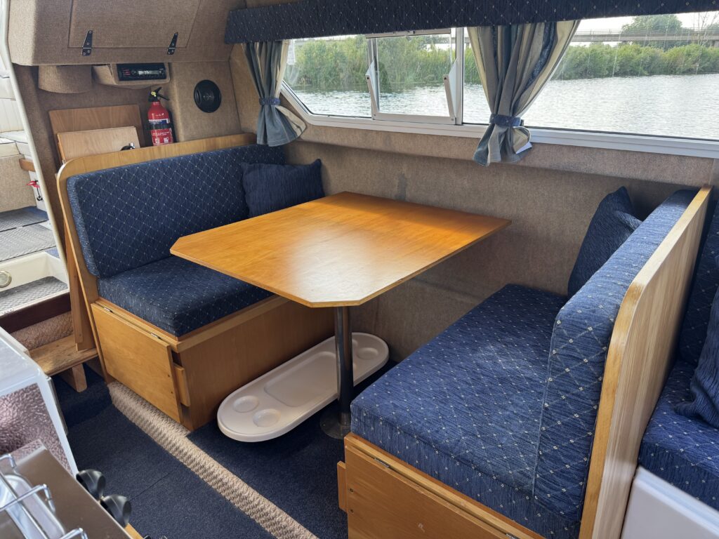 An interior seating area in a boat, featuring a wooden table and two blue upholstered benches with backrests on either side. The area is well-lit by large windows that offer a view of the water and greenery outside.