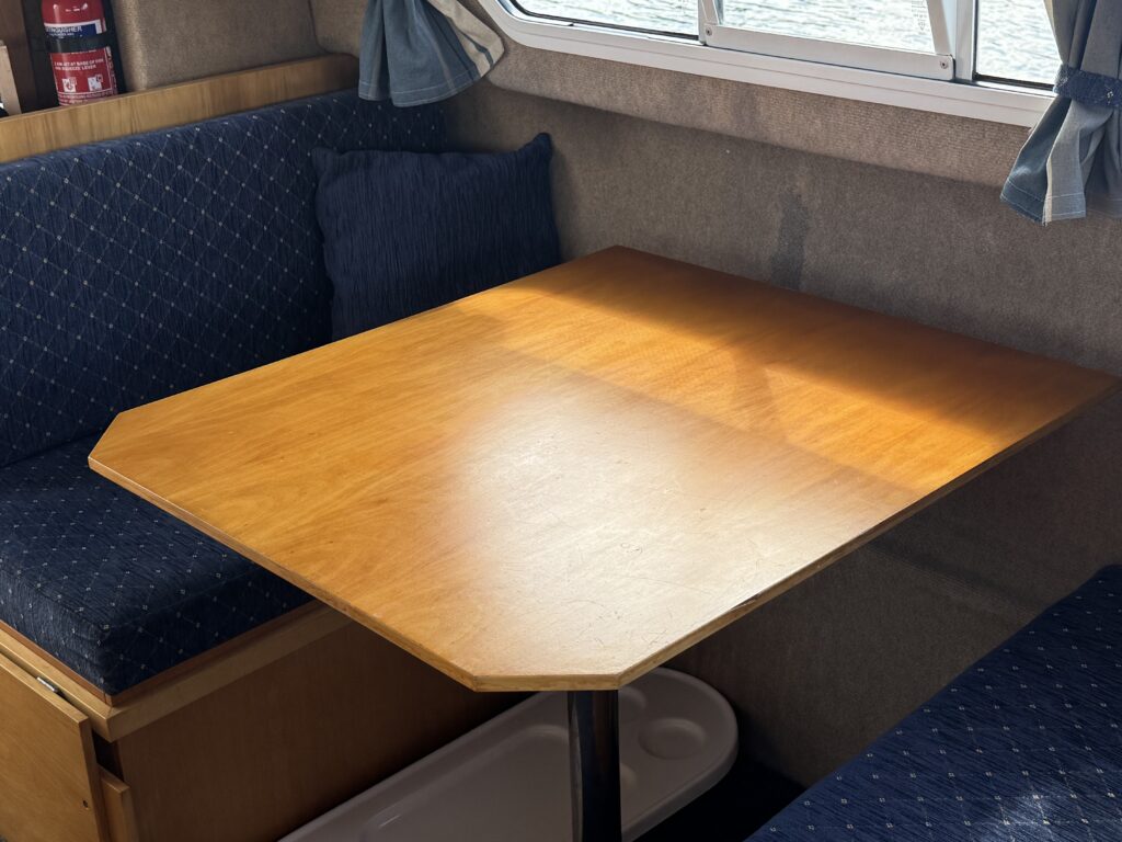 A wooden table with a light finish is placed in a compact seating area. The table is octagonal and sits on a metal pedestal, surrounded by blue upholstered benches with patterned cushions. Natural light streams in through a nearby window.