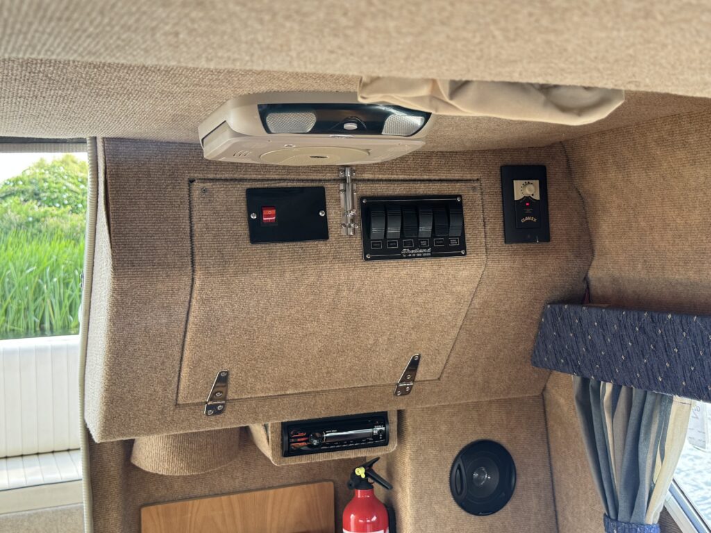 The interior of a vehicle featuring a light beige wall panel with various controls, including a light switch and multiple circuit breakers. A fire extinguisher is mounted on the lower part of the panel, with a round speaker visible to the right.