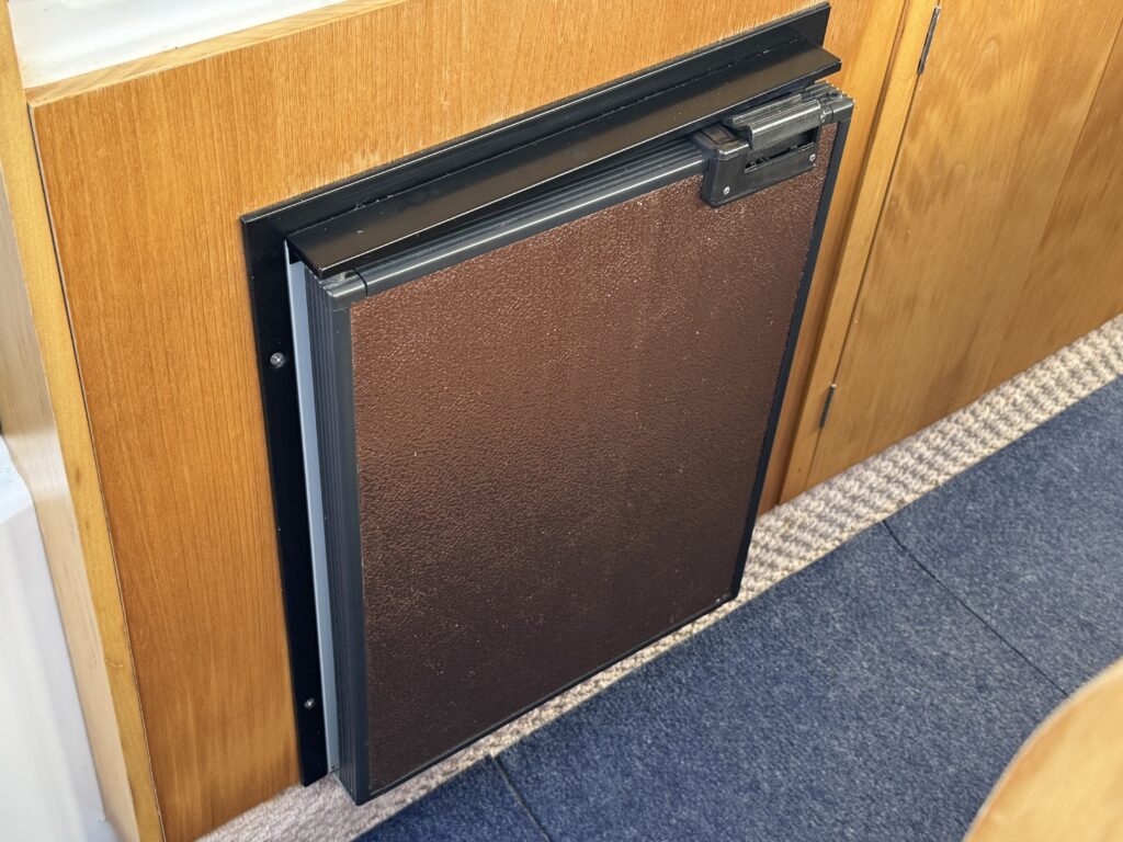 A dark brown storage compartment with a textured surface, set into a wooden wall. The compartment features a sliding mechanism with a metal handle, mounted beneath a smooth black frame and adjacent to a beige carpeted floor.