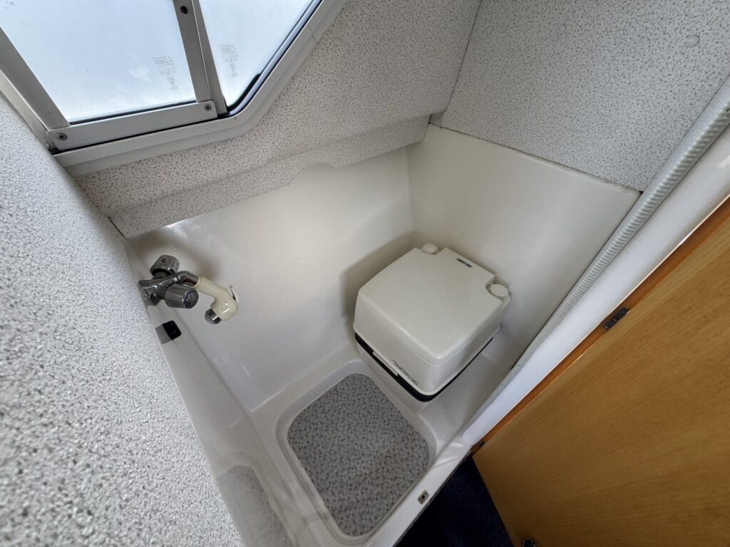 An interior view of a compact restroom showing a white toilet with a black seat located on a textured floor. A small window is visible above the toilet, and walls are covered in a light-colored, textured material.