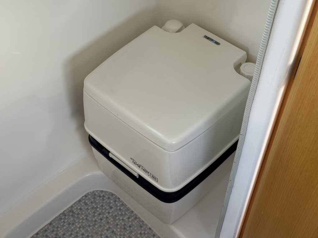A compact white portable toilet installed in a small bathroom. The toilet features a top lid and is positioned on a textured gray floor, with a wooden wall partially visible in the background.