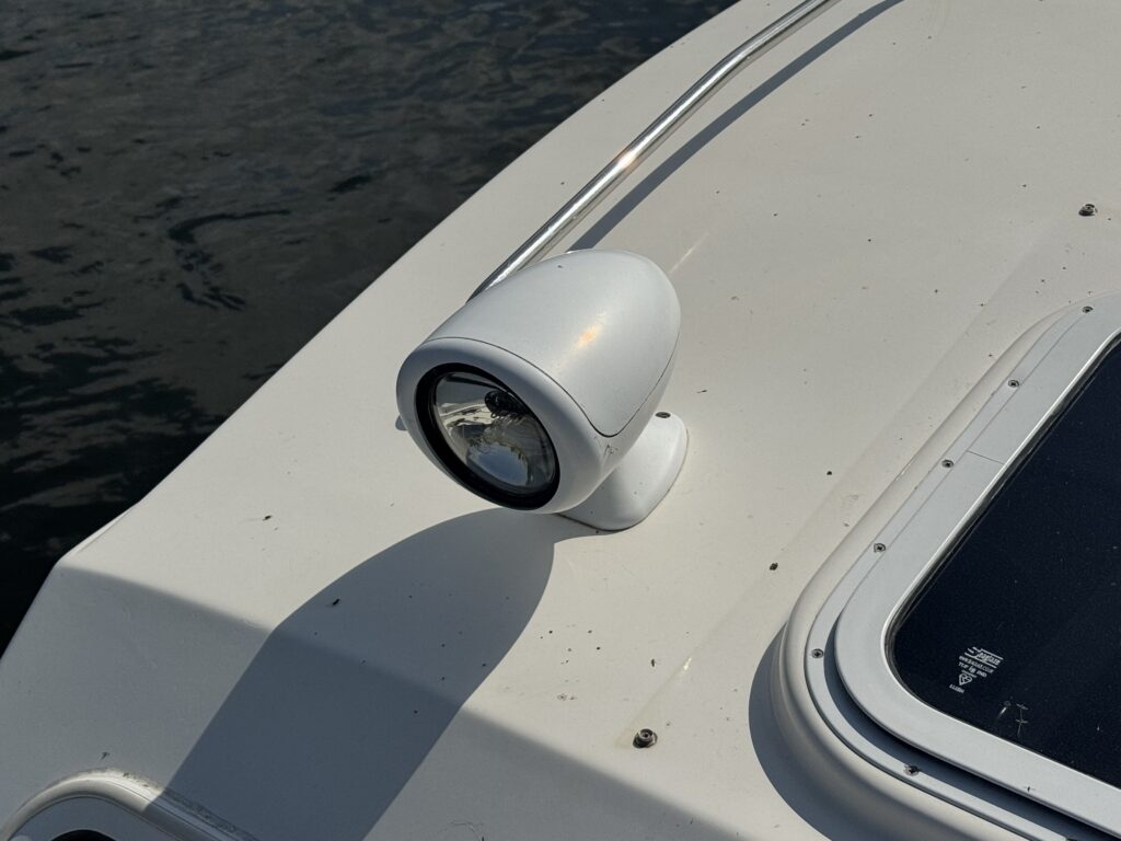 A close-up view of a white spotlight mounted on the deck of a boat. The spotlight is positioned near a railing, with the water visible in the background.