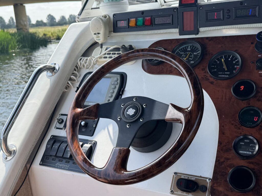 A close-up view of a boat's steering wheel made of polished wood and metal, positioned against a dashboard with various dials and controls. The background shows a waterway with greenery on either side and a bridge in the distance.