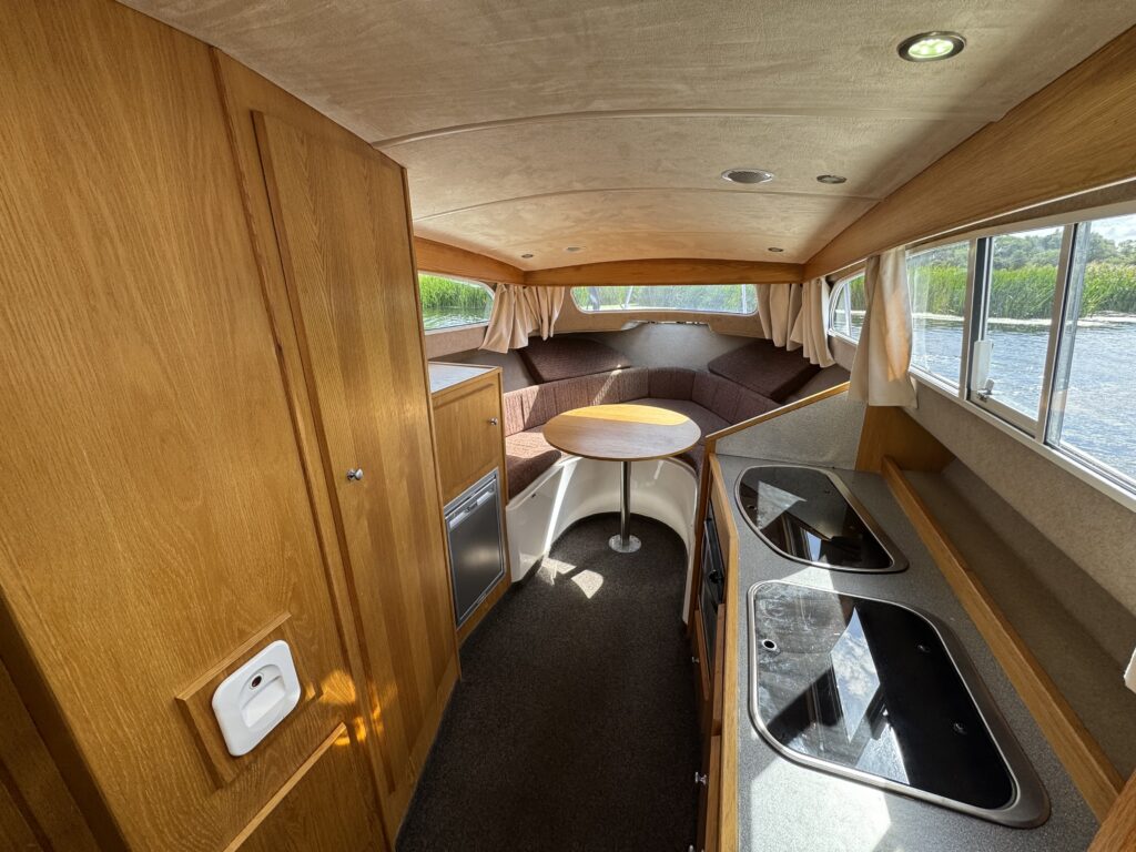 The interior of a boat features wooden cabinetry and countertops. On the right, there are two black sinks; a circular table is at the center, surrounded by cushioned seating. Large windows provide natural light, and there are curtains drawn back.