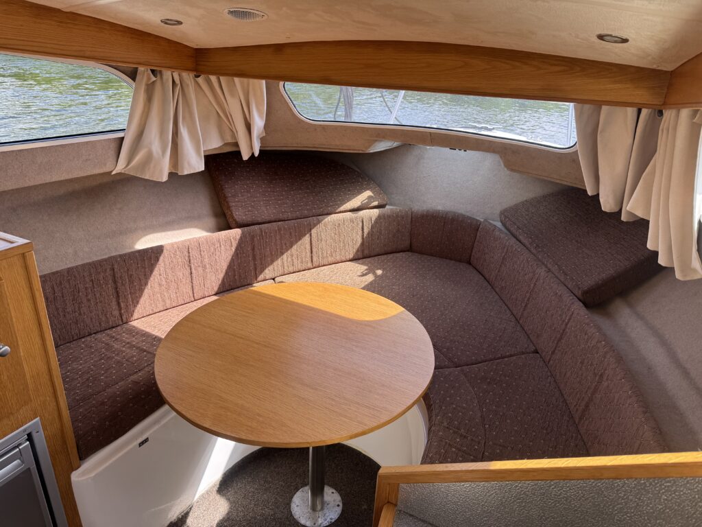 The interior of a small boat features a seating area with curved brown cushions surrounding a round wooden table. Sunlight streams in through a window, illuminating the cozy space.