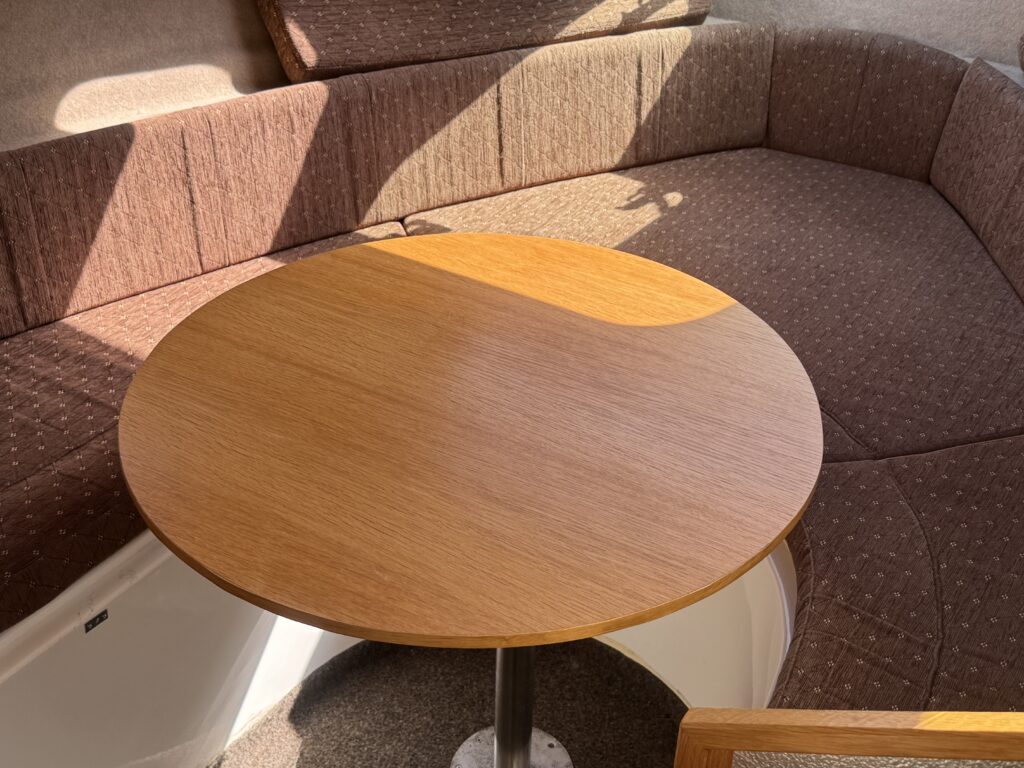 A round wooden table with a smooth surface is positioned in the center of a cozy seating area. The surrounding seating features a brown upholstered cushion that follows the curve of the space, with natural light illuminating the scene.