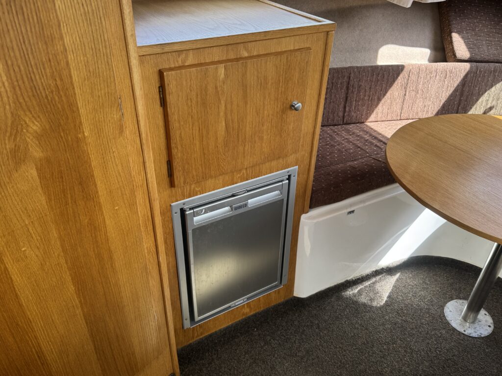 An interior view of a boat showing a wooden cabinet with a small refrigerator mounted below. The furniture includes a round table and a cushioned seating area with brown upholstery, illuminated by natural light.