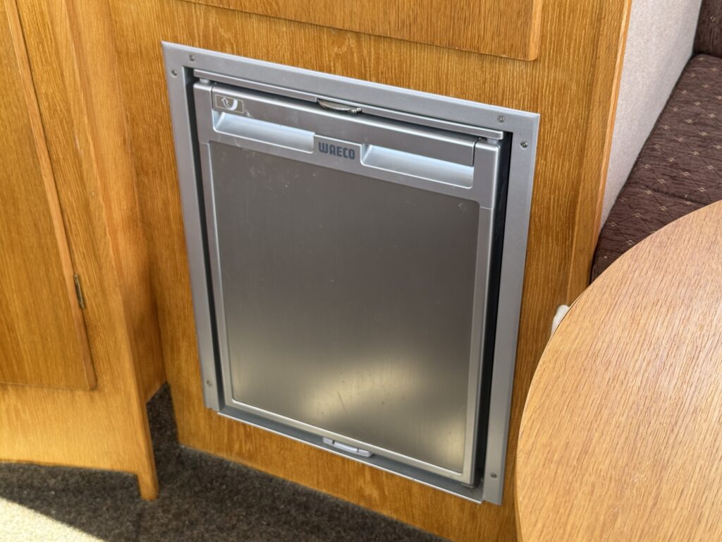 A compact refrigerator with a silver finish, built into a wooden cabinet. The refrigerator features a flush door design and is located next to a round wooden table in a cozy interior space.