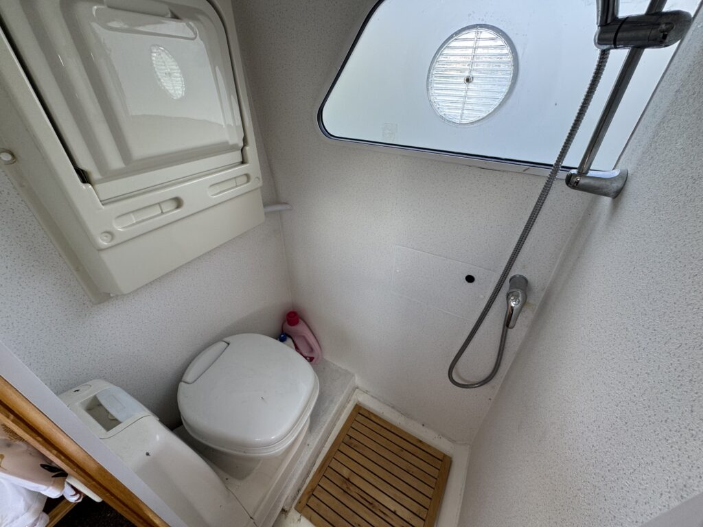 A compact bathroom featuring a white toilet, a shower head mounted on the wall, and a small wooden mat on the floor. A round window is visible on one wall, providing natural light, while a small storage compartment is installed above the toilet.