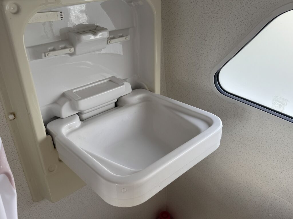 A foldable white sink is mounted on a wall, with its basin opened for use. The background shows a small window with a view of outside, and the interior features light-colored walls.